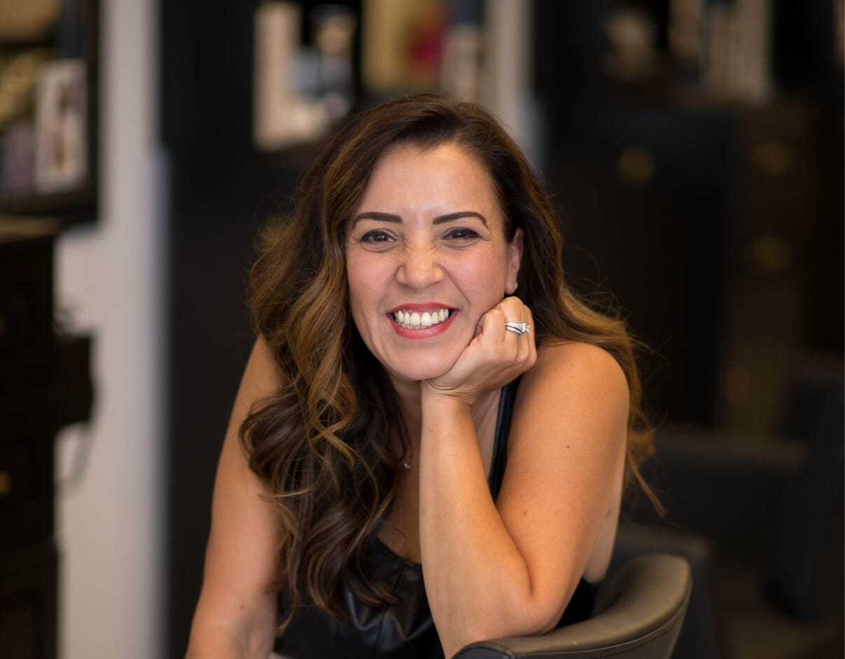 Smiling woman seated in a salon, with hand resting on chin.