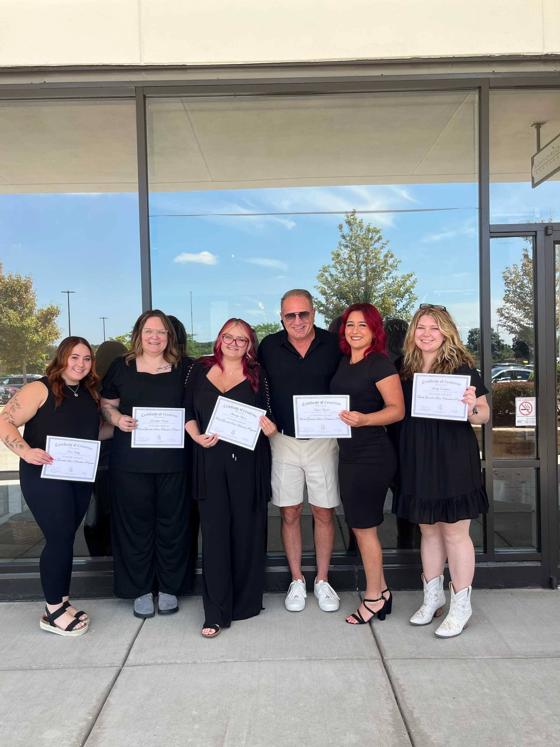 Group of six people holding certificates, standing in front of a building.