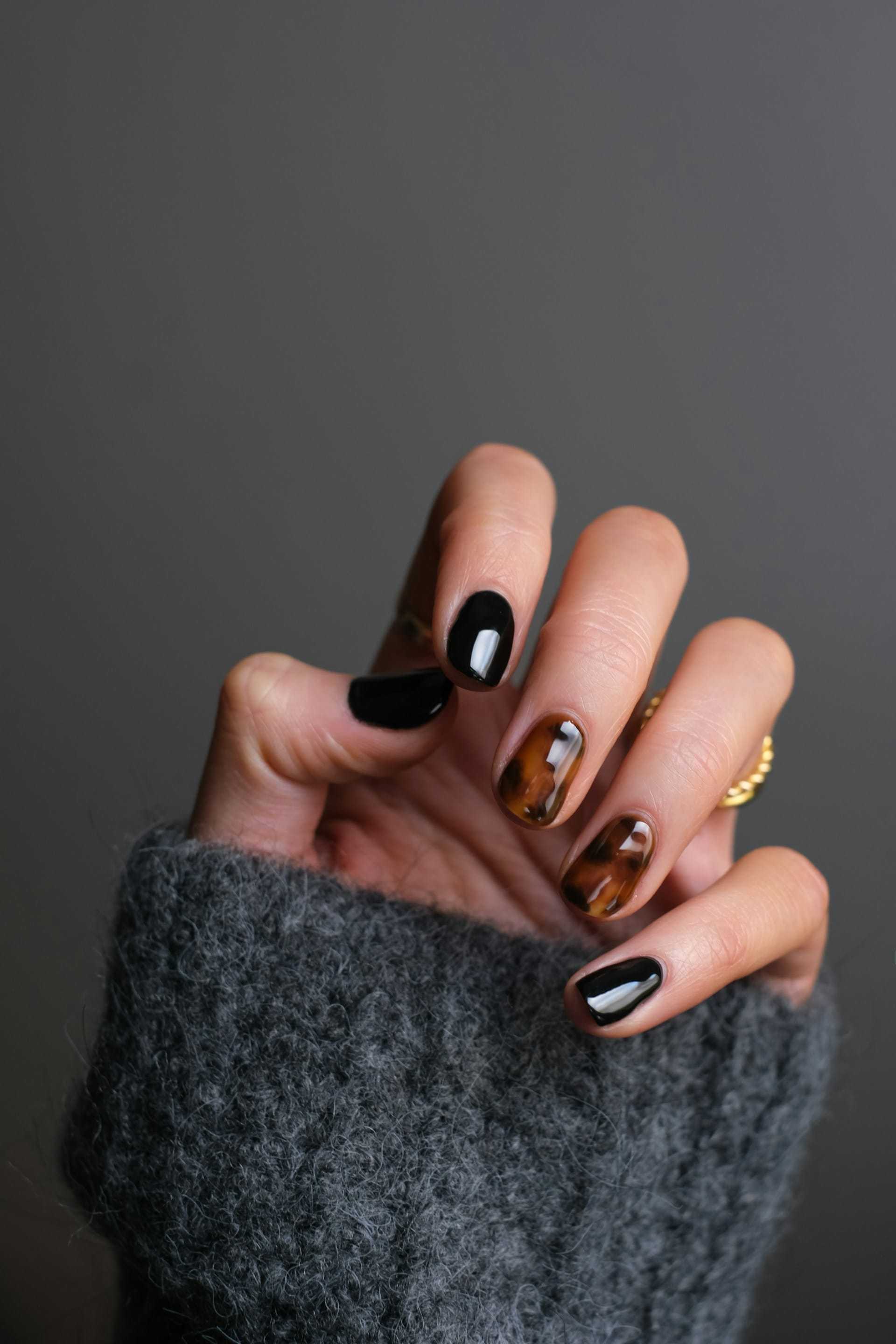 Hand with black and tortoiseshell nails, gray sweater, and gold rings.